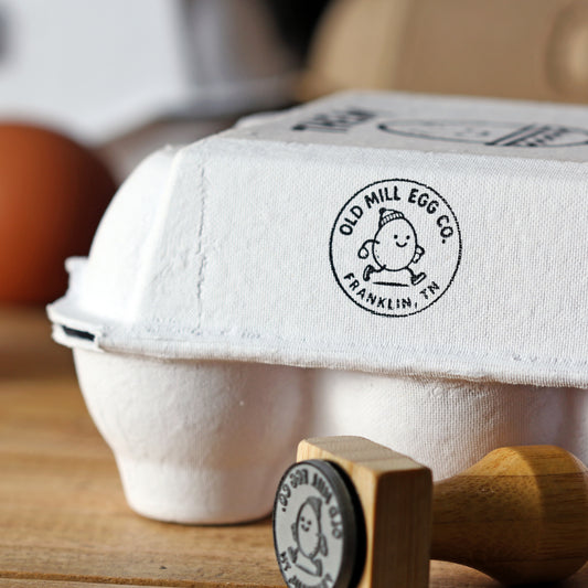 egg carton logo stamp on white pulp carton; small round badge; black ink; wood-handled stamp shown.