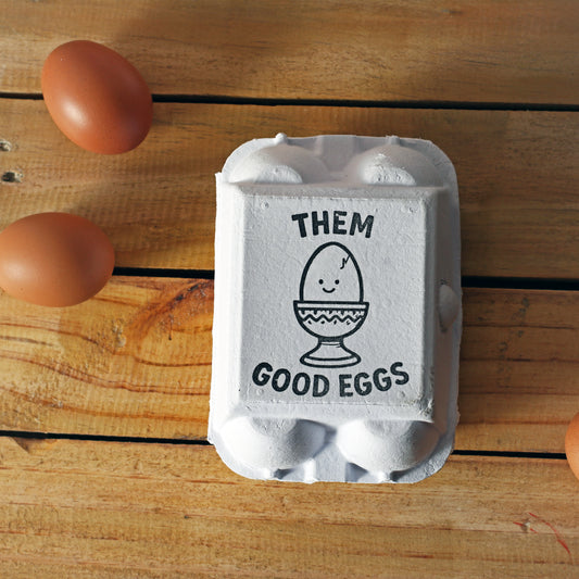 "Them Good Eggs" phrase with smiling egg icon stamped on small white egg carton; black ink; lid branding.